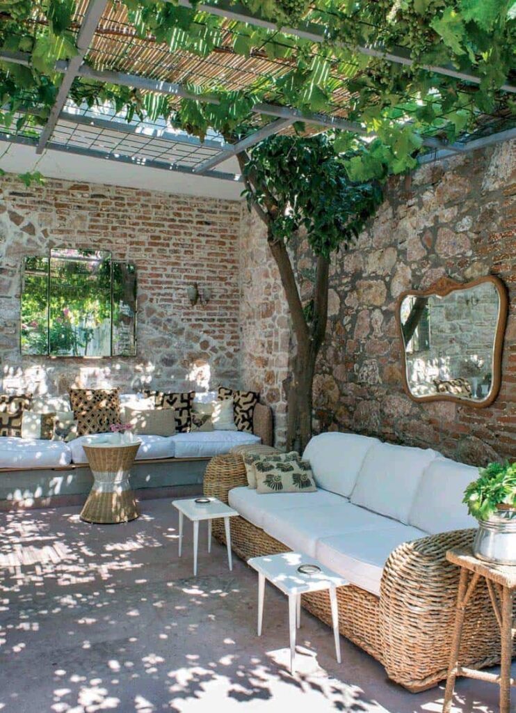 A beautiful shaded patio with ratan sofas with white cushions and neutral shades of throw pillows, natural stone walls, a tree, a vine, and a plant.